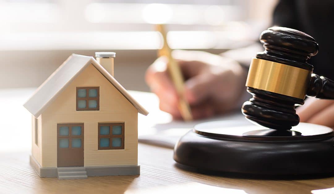 Ein Mann unterschreibt ein Dokument auf einem Tisch und im Vordergrund stehen ein Modellhaus und ein Richterhammer | Immobilie in der Scheidung